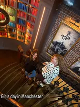 A photo of two women posing on a staircase in a venue with a library-themed interior, featuring a portrait of Winston Churchill. The image captures a social outing, likely at a bar or pub, with text overlay indicating a 'Girls day in Manchester.'