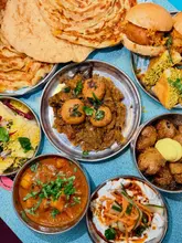 A high-angle, overhead flat-lay shot of a vibrant Indian street food feast, featuring various bowls of curries, chaat, sliders, and flatbreads arranged on a blue table. The lighting is bright and even, highlighting the textures and colors of the dishes, creating an inviting and communal dining atmosphere.