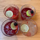 A top-down, well-lit photograph of four different margaritas arranged on a wooden table. The drinks are garnished with lime wheels, cherries, and strawberries, with some featuring a chili-salt rim, presenting a vibrant and inviting selection of cocktails.