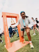 A candid, high-energy photograph capturing a runner celebrating at a 'PB Bell' station during a running event. The image effectively conveys the community spirit and excitement of the occasion, with the subject's genuine expression serving as the focal point.