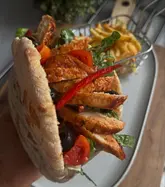 A high-quality, appetizing close-up shot of a homemade chicken pitta sandwich held in the hand, with a side of fries in a wire basket in the background. The lighting is soft and natural, highlighting the textures of the grilled chicken and fresh salad ingredients.