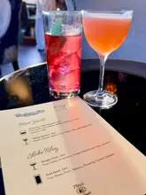 A close-up, shallow depth-of-field shot focusing on a cocktail menu from a bar takeover, with two cocktails blurred in the background. The image highlights the menu offerings rather than the drinks themselves.