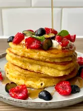 A high-quality, close-up shot of a stack of pancakes being drizzled with syrup, garnished with fresh raspberries, blueberries, and cream cheese. The image is bright and well-composed, focusing on the texture of the pancakes and the action of the syrup pour.