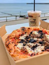 A close-up shot of a pizza topped with haggis and black pudding in a cardboard box, positioned in the foreground with an iced coffee cup behind it, set against a blurred seaside background.