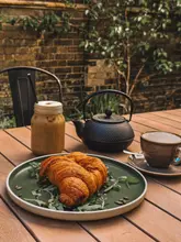 A high-quality, well-composed shot of a cafe table featuring a flaky croissant served on a bed of greens, accompanied by an iced coffee and a cast-iron teapot. The setting is an outdoor courtyard with a rustic brick wall background, evoking a relaxed, pleasant atmosphere.