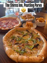 A high-angle, slightly cluttered table spread shot featuring two pizzas, loaded fries, onion rings, and halloumi, with a text overlay identifying the venue.