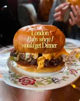 A close-up shot of a pub-style burger on a floral plate, used as a cover image for a listicle about London pubs. The image focuses on the burger, which appears juicy and well-prepared, though the composition is slightly compromised by the inclusion of the creator's hand and background distractions.