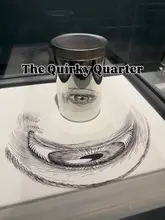 A clean, well-lit photograph of an optical illusion exhibit at The Quirky Quarter, featuring a cylindrical tin placed on a surface with a matching drawing to create a cohesive visual effect.