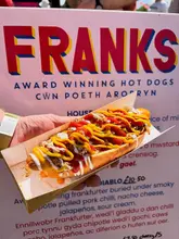 A close-up, eye-level shot of a loaded hot dog held in a cardboard tray, captured at an outdoor food festival with a menu board visible in the background.