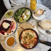 A bright, well-composed flat-lay photograph of a brunch spread featuring shakshuka, avocado toast with poached eggs, and berry-topped French toast, accompanied by coffee and juice on a marble table.