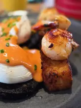 A close-up, shallow depth-of-field shot of a 'fisherman's benedict' dish featuring seared scallops, a hash brown, poached egg, and black pudding, served at a marina-side restaurant.