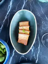 A high-quality, overhead shot of a portion of crispy pork belly served in a dark, textured bowl on a black marble table. The lighting is even and highlights the texture of the meat, making it look appealing.