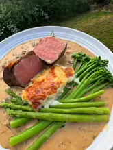 A high-quality, overhead food photograph showcasing a home-cooked meal consisting of a medium-rare fillet steak, potato gratin, tenderstem broccoli, and asparagus, all served in a rich sauce.