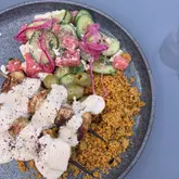 An overhead shot of a plate featuring chicken skewers covered in a thick white sauce, served with couscous and a watermelon, cucumber, and feta salad.