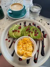 A top-down shot of a brunch meal consisting of avocado toast topped with pomegranate seeds and a side of scrambled eggs, accompanied by a cup of coffee and a glass of water, served on a speckled table at a cafe.