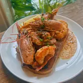 A close-up, slightly elevated angle shot of a dish featuring prawns served on a slice of toasted bread, likely sourdough, with a lemon wedge on the side. The lighting is natural and soft, highlighting the texture of the prawns and the sauce.