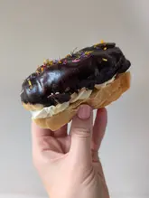 A high-quality, close-up shot of a chocolate eclair being held by a hand against a plain, neutral background. The eclair is topped with edible flowers and filled with cream, presented in a simple, effective style that highlights the texture and quality of the pastry.