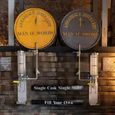 A symmetrical, high-quality shot of the 'Fill Your Own' whisky station at Annandale Distillery, featuring two casks labeled 'Man O' Words' and 'Man O' Sword' mounted above industrial-style filling mechanisms. The rustic wooden backdrop and clear, professional lighting create an inviting and authentic atmosphere for whisky enthusiasts.