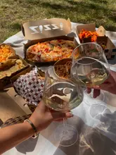A first-person perspective shot of a picnic spread featuring various takeaway boxes from Zizzi, including pizza, pasta, and sides, accompanied by two glasses of white wine being toasted.