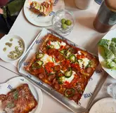 A top-down flat-lay photograph of a Detroit-style pizza, a side salad, and a small bowl of olives on a table with branded paper. The image captures a casual dining experience with a focus on the textures of the food.