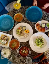 A high-quality, overhead flat-lay shot of a generous spread of Indian dishes, including curries, rice, naan, and a cocktail, arranged on a dark, textured table. The lighting is soft and even, highlighting the textures and colors of the food.