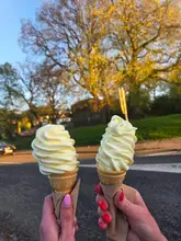 A first-person perspective shot of two soft-serve ice cream cones held up against a sunset background, capturing a casual, pleasant moment.