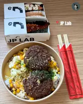 A top-down, well-lit photograph showcasing a takeaway meal from JP Lab. The image features a selection of sushi tacos in branded boxes alongside a hearty bowl of wagyu beef patty rice topped with egg. The composition is clean and focused, effectively highlighting the textures of the food.