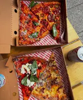 A high-quality, overhead flat-lay shot of four different pizza slices served in takeaway boxes on a wooden table. The lighting is natural and bright, highlighting the textures of the toppings and the crispy crusts, making the food look highly appealing.
