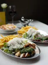 A close-up shot of two metal trays serving chicken with a green herb sauce, fries, and a side salad, with a bowl of mac and cheese and a glass of red wine visible in the background.