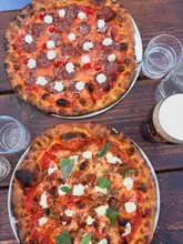 A high-quality, overhead flat-lay photograph of two pizzas served on a rustic wooden table, accompanied by glasses of water and a drink. The lighting is bright and natural, highlighting the textures of the crispy crusts and the fresh toppings. The composition is clean and inviting, effectively capturing the casual, social atmosphere of the venue.