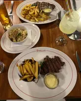 A top-down, overhead shot of a meal at The McMillan, featuring two plates of steak and chips, a side of mac and cheese, and a limoncello spritz. The image captures a casual dining experience, though the composition is somewhat cramped with cutlery cut off at the edges.
