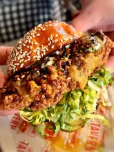 A high-quality, close-up shot of a crispy chicken burger being held by hand, effectively showcasing the texture of the fried chicken, the toasted sesame seed bun, and the fresh lettuce. The background is softly blurred, ensuring the focus remains entirely on the food, while the branded paper adds a nice touch of context.