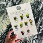 A clear, well-lit, and informative shot of a printed menu at Lulu's Coffee Shop, held by a customer. The image effectively showcases the variety of iced matcha drinks available, providing clear pricing and visual representations of the menu items against a stylish marble background.