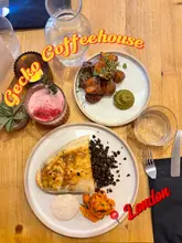 An overhead shot of a dinner spread at Gecko Coffeehouse, featuring a plate of Lebanese Arayes with yoghurt and carrots, and a side of crispy potatoes with green goddess dressing, alongside a pink mocktail.
