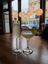 A static shot of two cocktails served in a flute and a coupe glass, placed on a wooden table in a bar setting with a view of the street through a window.