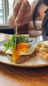 A montage-style reel showcasing a brunch experience at a riverside venue in Manchester. The video features close-ups of steak and eggs, bagels, coffee, and mimosas, interspersed with shots of the restaurant's interior and outdoor seating area.