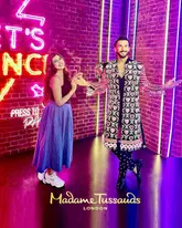 A photo of a creator posing with a wax figure of Ranveer Singh at Madame Tussauds London, set against a neon-lit brick wall background.