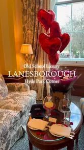 A promotional shot of a luxury hotel room at The Lanesborough, featuring a celebratory setup with red heart-shaped balloons and small plates under cloches on a coffee table. The image captures an elegant, regal interior aesthetic suitable for a high-end staycation.