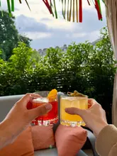 A first-person perspective shot of two people clinking cocktail glasses together on an outdoor terrace, with lush greenery in the background and soft, natural sunlight filtering through.