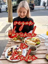 A bright, outdoor brunch scene featuring a woman smiling at a table laden with vegan dishes, including waffles and shakshuka, set against a street-side backdrop in London.