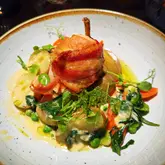A top-down, well-lit photograph of a bacon-wrapped chicken ballotine served with potatoes, greens, and a creamy sauce. The image is sharp and clearly captures the textures of the dish, though the plating is somewhat rustic and cluttered.