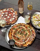 A high-quality, overhead flat-lay shot of a table spread featuring two pizzas, a side dish, a bottle of rosé, and various drinks, set on a rustic wooden table.