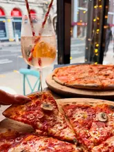 A close-up shot of a hand holding a slice of pizza, with a cocktail in the foreground and a whole pizza on a wooden board in the background. The setting appears to be a casual bar or restaurant with a view of the street outside.