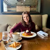 A portrait-style shot of a woman enjoying a brunch spread featuring pancakes, a coffee, and a croissant at a restaurant.