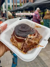 A close-up, handheld shot of a Tiramisu-flavoured cinnamon bun served in a white takeaway container, captured at an outdoor market.