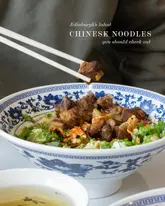 A high-quality, close-up shot of a bowl of Lanzhou-style hand-pulled beef noodles, featuring a pair of chopsticks lifting a piece of tender braised beef. The image is well-composed, highlighting the texture of the meat and the vibrant colors of the broth and garnishes, set against a traditional blue and white patterned bowl.