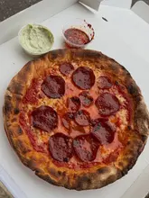 A top-down, overhead shot of a homemade pepperoni pizza served in a cardboard delivery box, accompanied by two small plastic tubs of sauce.