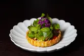 A high-quality, professional food photograph featuring an intricate, beautifully plated tartlet garnished with fresh herbs and edible flowers, set against a dark, moody background that emphasizes the dish's vibrant colors and textures.