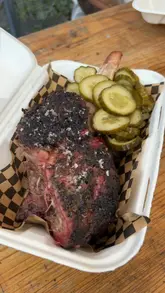 A food review video featuring a creator trying various smoked meat dishes, including short rib, brisket, a brisket burger, dirty fries, and mac and cheese triangles at a BBQ venue.