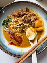 A top-down, well-lit photograph of a bowl of curry laksa, featuring crispy chicken, a boiled egg, and bean sprouts, served with chopsticks and a spoon on a clean surface.
